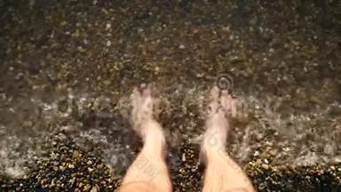 一个赤脚的人站在水中的鹅卵石海岸上。 照相机的焦点在那个人`<strong>脚上</strong>。